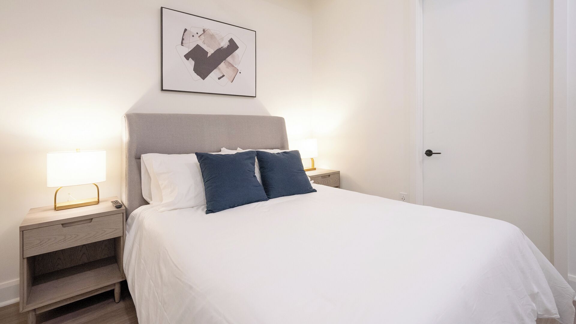 A neatly made bed with white bedding and two blue pillows, flanked by bedside tables with lamps, in a room with a plain white door and a framed abstract artwork on the wall.