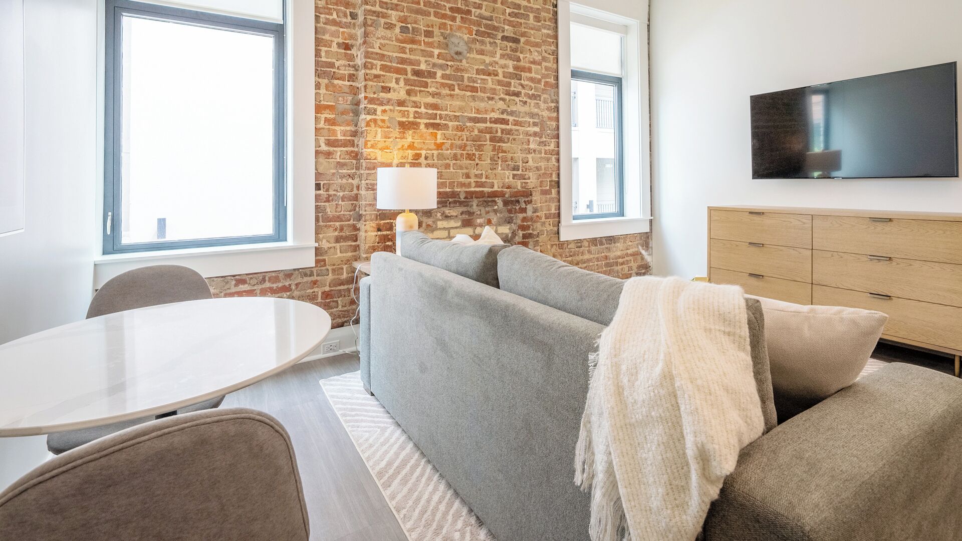 A modern living room with a brick wall, a grey sofa, a white table, and a TV mounted on the wall.