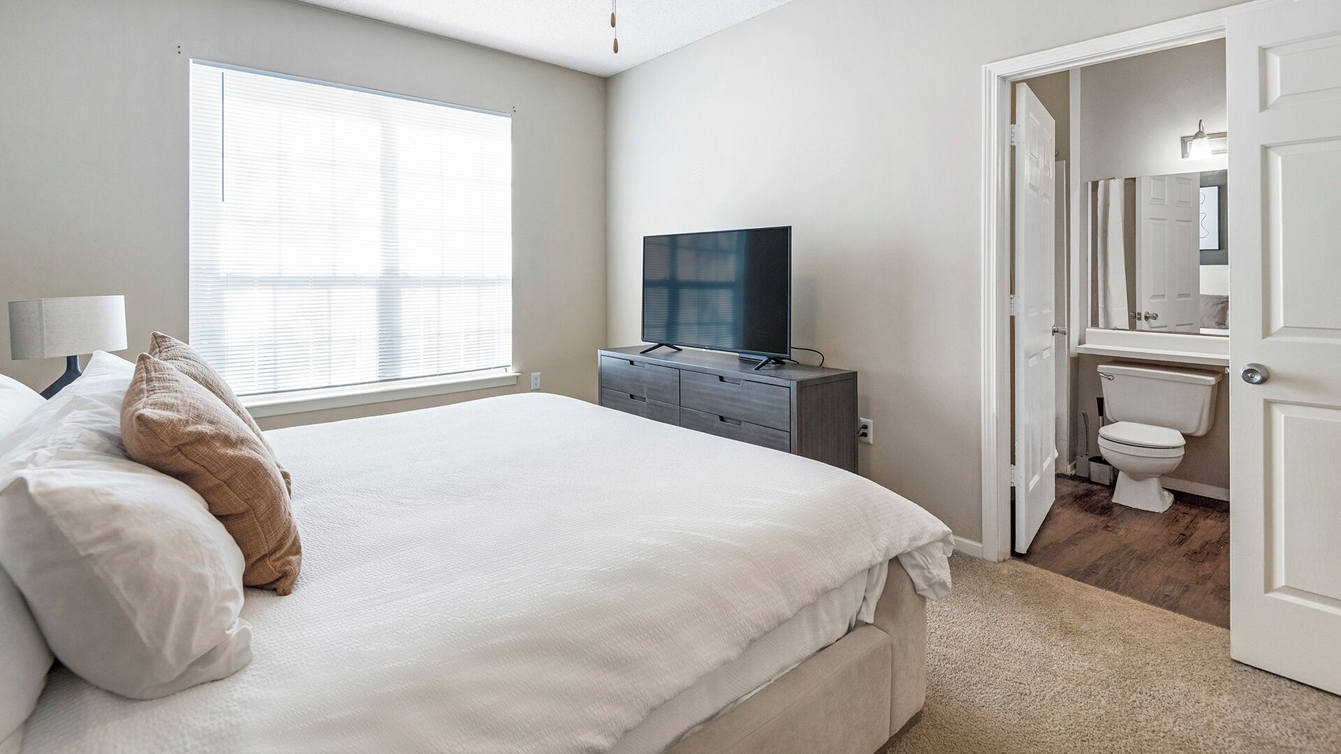A bedroom with a bed, a TV on a dresser, a window, and a bathroom visible through an open door.