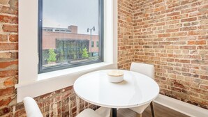 Ein Zimmer mit Backsteinwand, einem runden weißen Tisch mit einer Holzschüssel und einem Fenster mit Blick auf ein Gebäude mit Bäumen.