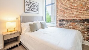 Ein ordentlich bezogenes Bett mit weißen Leinen und grauen Kissen in einem Zimmer mit Backsteinwand und Fenster.