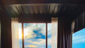 Ein Zimmer mit großem Fenster und Blick auf ein Gewässer, zwei blaue Stühle, ein kleiner Holztisch und eine Vase mit Blumen.