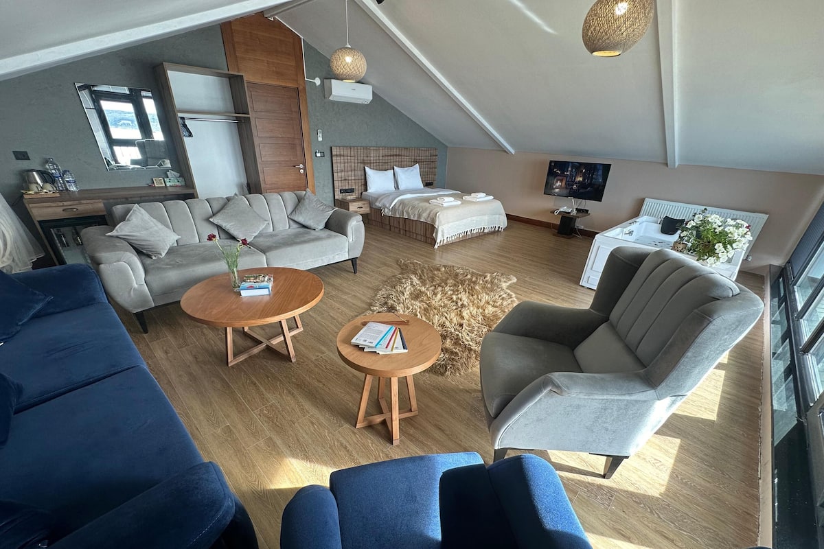 A modern hotel room with a loft-style layout, including a living area with sofas, a dining table, and a bedroom with a large bed and a TV.