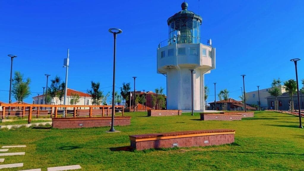 A lighthouse with a cylindrical base and a glass lantern room, surrounded by a grassy area with pathways and benches.