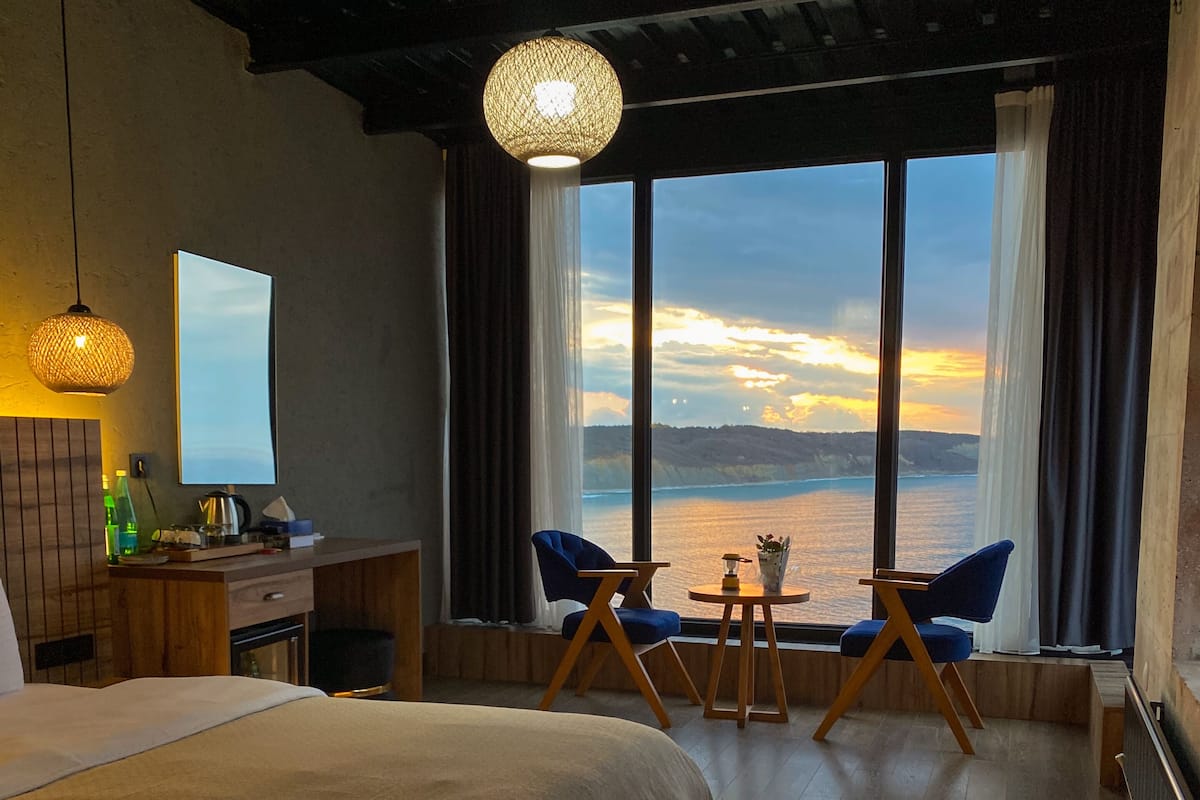 A hotel room with a large bed, a desk with a chair, a small table with two chairs, and a view of the sea through large windows.