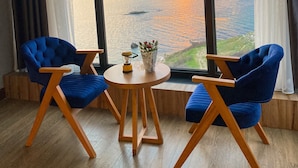 Ein Zimmer mit zwei blauen Stühlen, einem kleinen Holztisch und Meerblick bei Sonnenuntergang durch große Fenster.