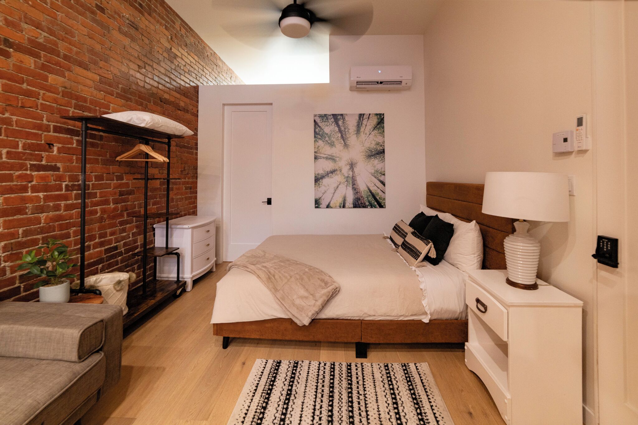 A bedroom with a brick wall, a bed with a wooden headboard, a bedside table with a lamp, a hanging wardrobe, and a potted plant.