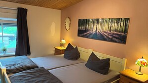 A bedroom with two beds, a nightstand with a lamp, a wall clock, and a forest path painting. - Ferienhaus "stück - Davon " Nr. 2 - Ruhig, Garten, Grillplatz, See, Schorfheide (Joachimsthal)