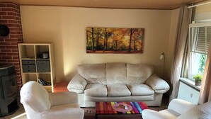 A living room with a beige sofa, two white armchairs, a colorful rug, a painting of a forest, a brick wall, and a fireplace. - Ferienhaus "stück - Davon " Nr. 2 - Ruhig, Garten, Grillplatz, See, Schorfheide (Joachimsthal)