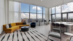 Un salon moderne comprenant un canapé, une table basse et des fauteuils, un coin cuisine avec un bar, et de grandes fenêtres offrant une vue sur la ville.