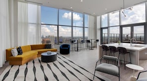 A modern living room with a sofa, coffee table, and chairs, a kitchen area with a bar, and large windows offering a city view.