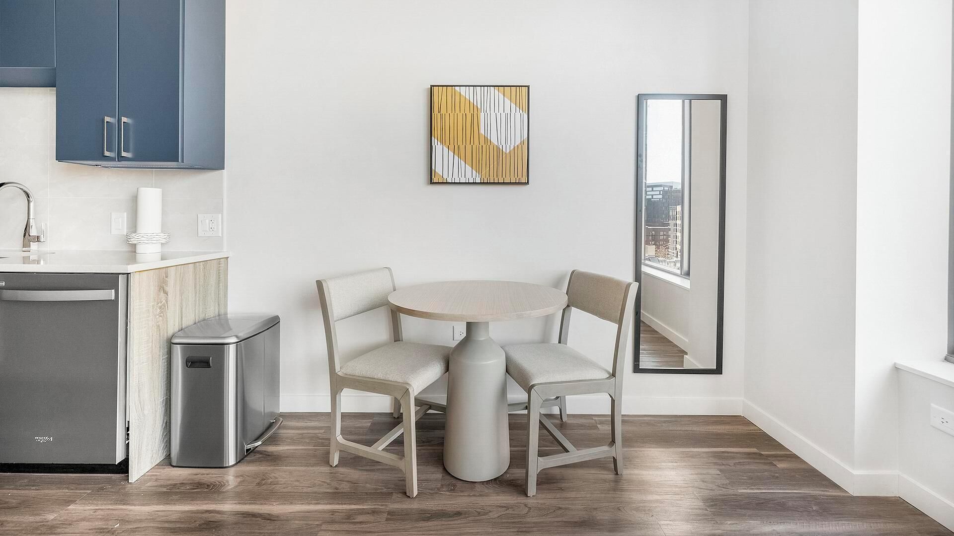 Een moderne keuken met een eethoek, met een ronde tafel en stoelen, een kleine prullenbak en een groot raam met uitzicht op de stad.