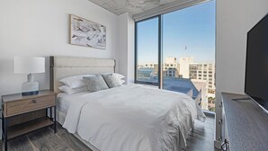 Ein modernes Hotelzimmer mit einem großen Bett, einem Nachttisch mit Lampe, einem Fernseher und einem großen Fenster mit Blick auf die Stadt.