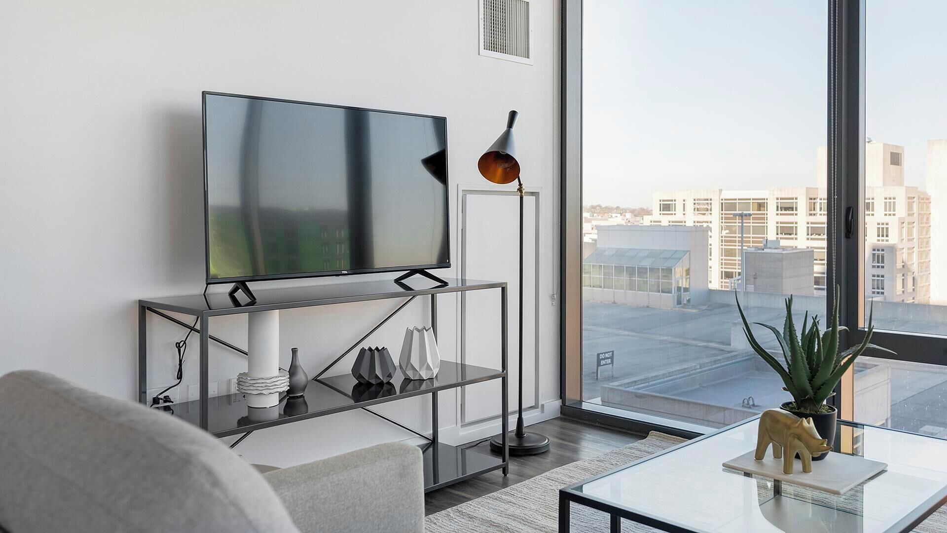 A modern living room with a large flat-screen TV, a floor lamp, and a view of the city through large windows.