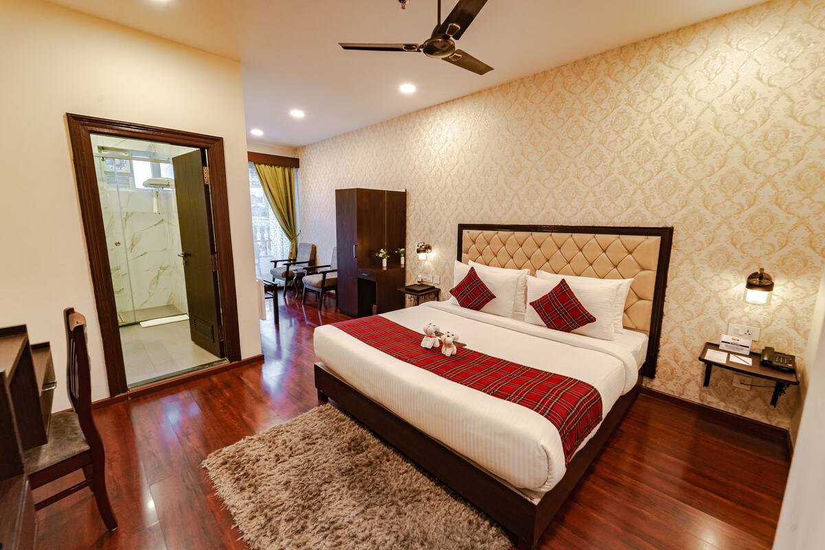 A bedroom with a large bed, wooden floor, a ceiling fan, a nightstand with a phone, and a door leading to a bathroom.