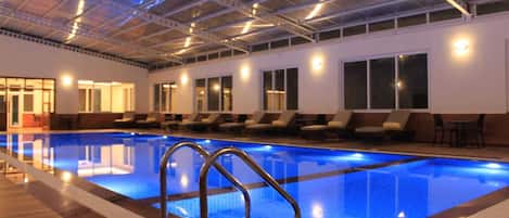 An indoor swimming pool with clear blue water, surrounded by lounge chairs and a high ceiling with exposed beams.