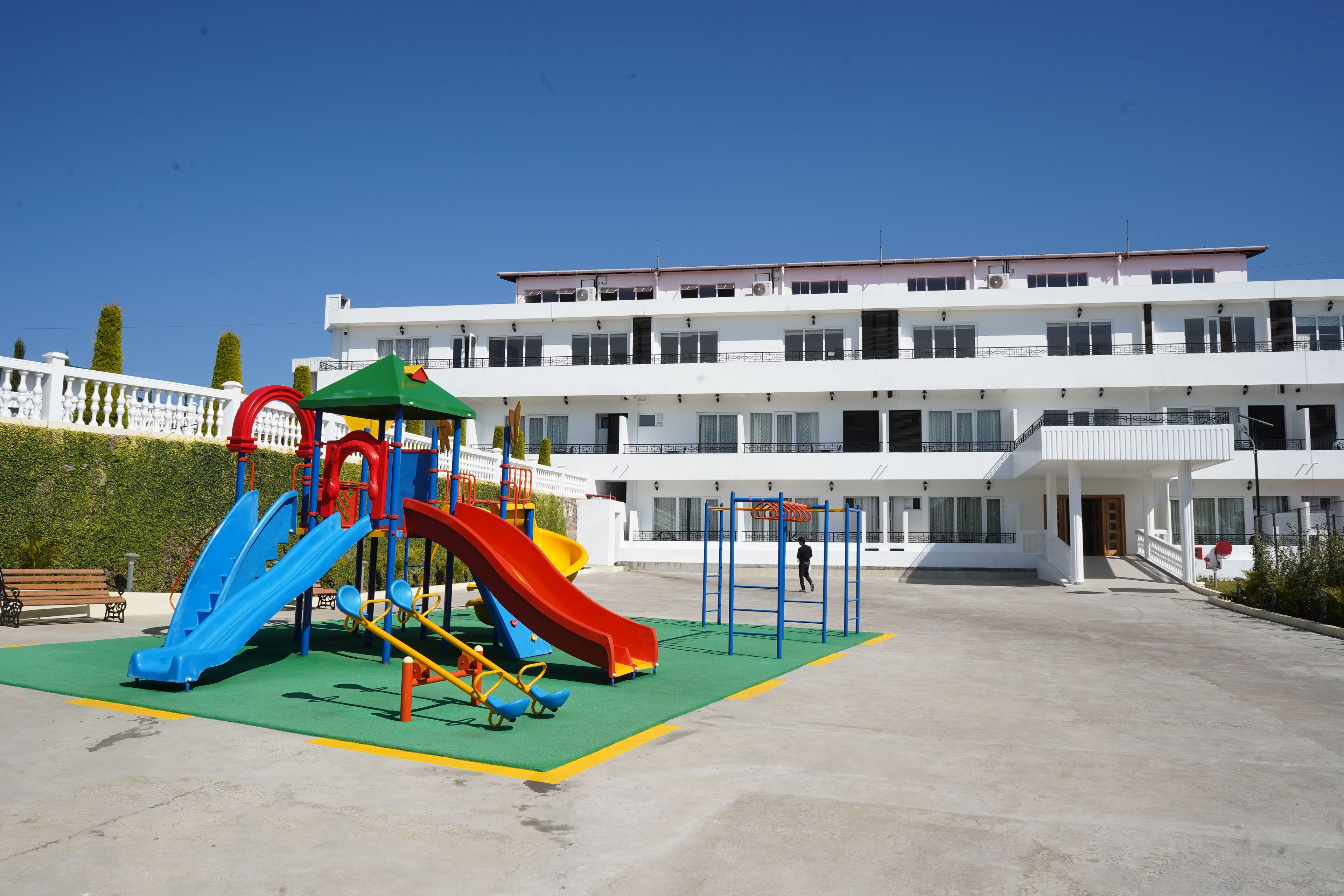 Un parque infantil con toboganes y columpios, un edificio blanco de varios pisos con balcones, y un cielo azul despejado.