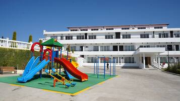 Un parque infantil con toboganes y columpios, un edificio blanco de varios pisos con balcones, y un cielo azul despejado.