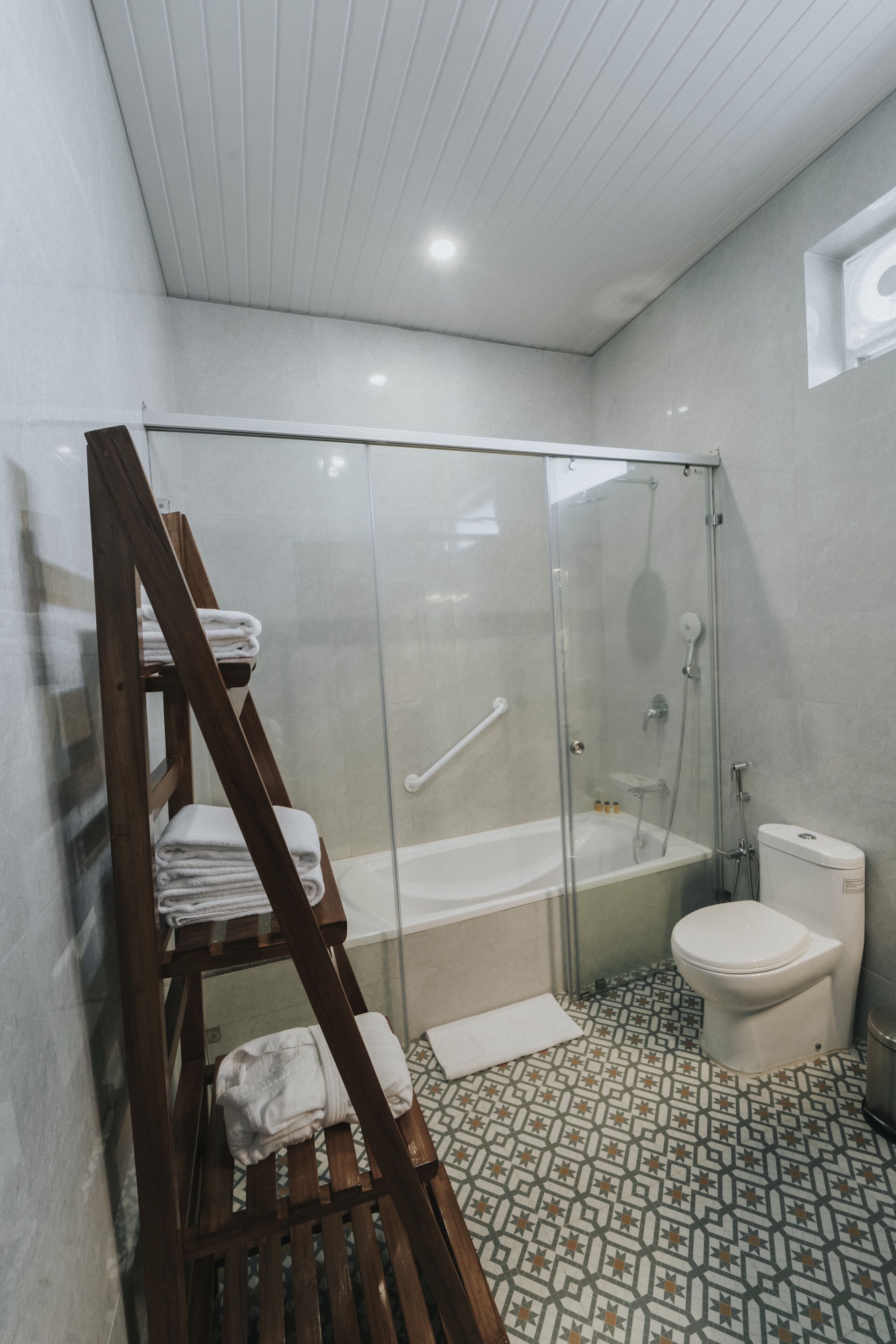 Et moderne badeværelse med en glasafskærmet brusekabine, en træhylde med håndklæder, et toilet og gulvfliser med et mønster.