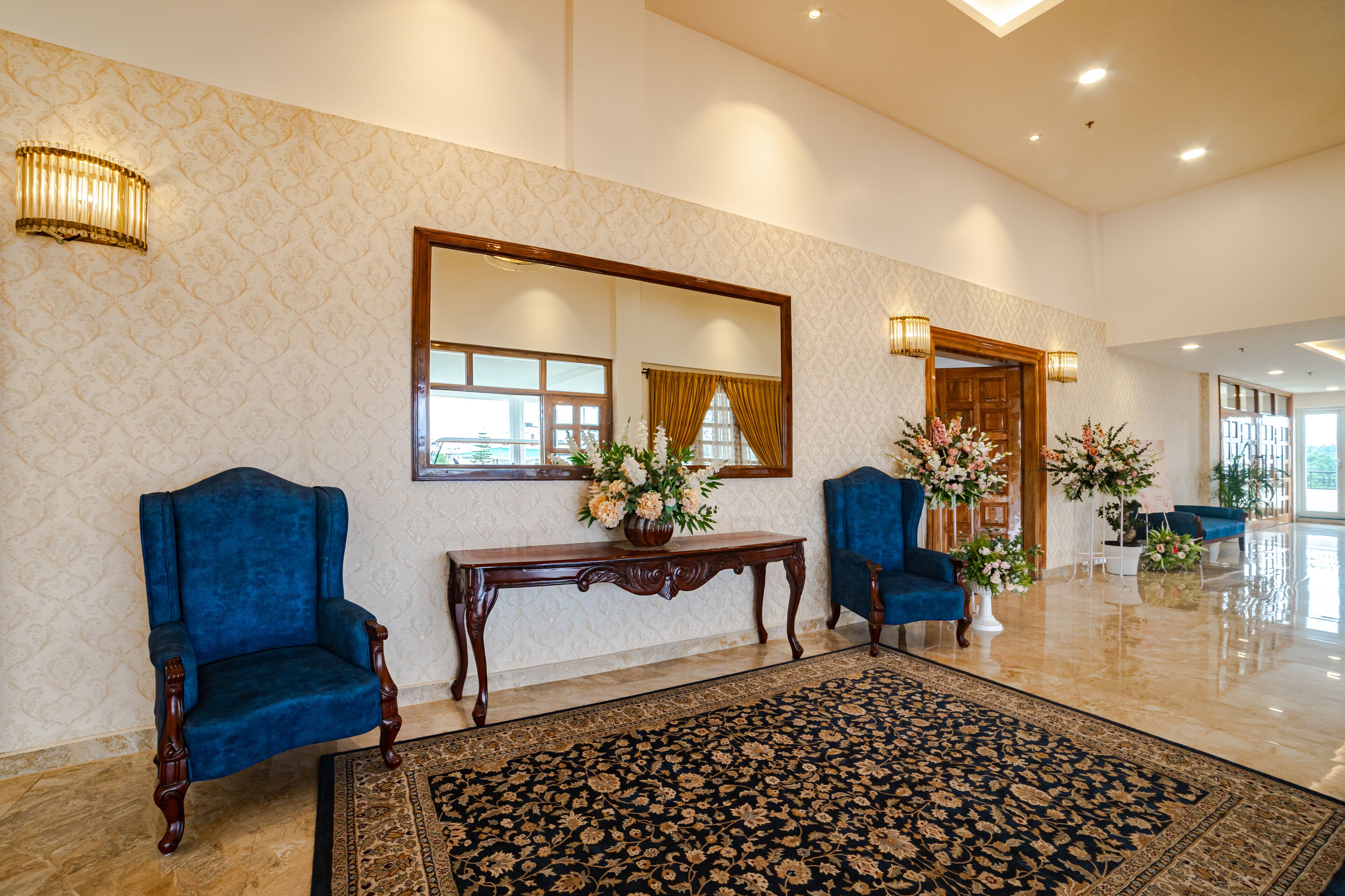 Um hall de entrada com duas poltronas azuis, uma mesa consola de madeira com um vaso de flores, um espelho grande e um tapete estampado.