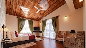 A bedroom with a wooden ceiling, a bed with white bedding, green curtains, a TV, and a floral armchair. - Windermere Hotel (Shillong)