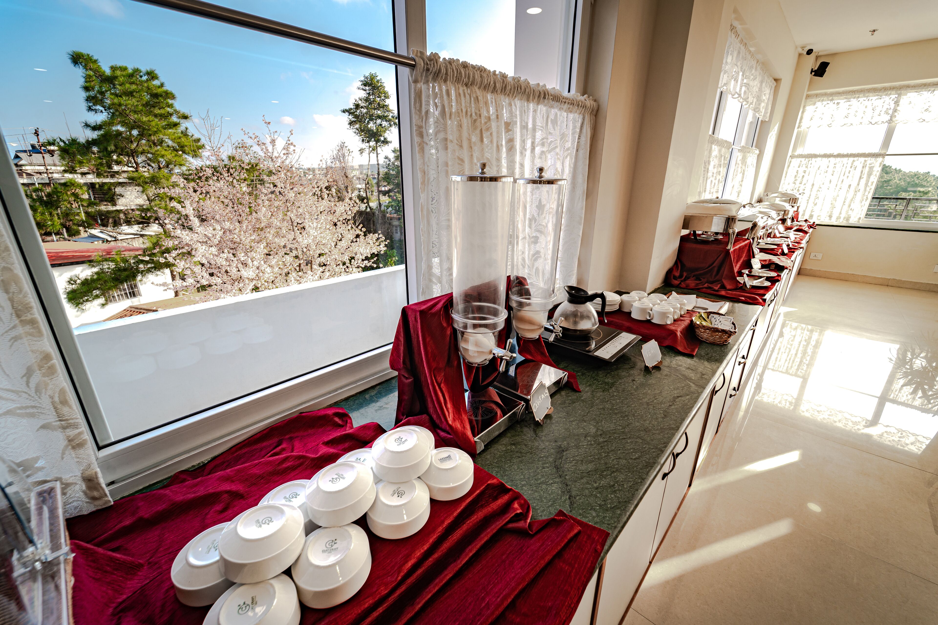 Une chambre d’hôtel avec une grande fenêtre, une table à manger dressée avec de l’assiette blanche et des nappes rouges, et une vue sur les arbres et les bâtiments à l’extérieur.