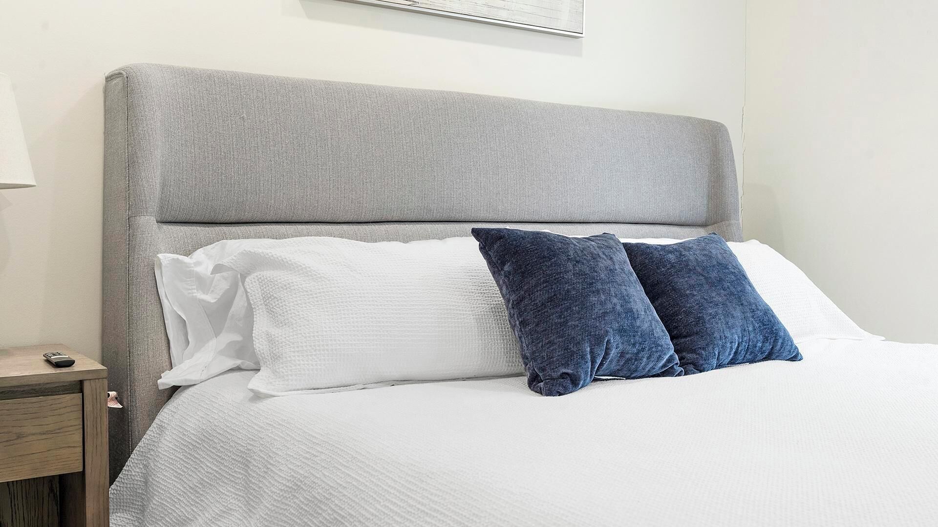 A bed with a grey upholstered headboard, white and blue pillows, and a wooden nightstand with a remote control.