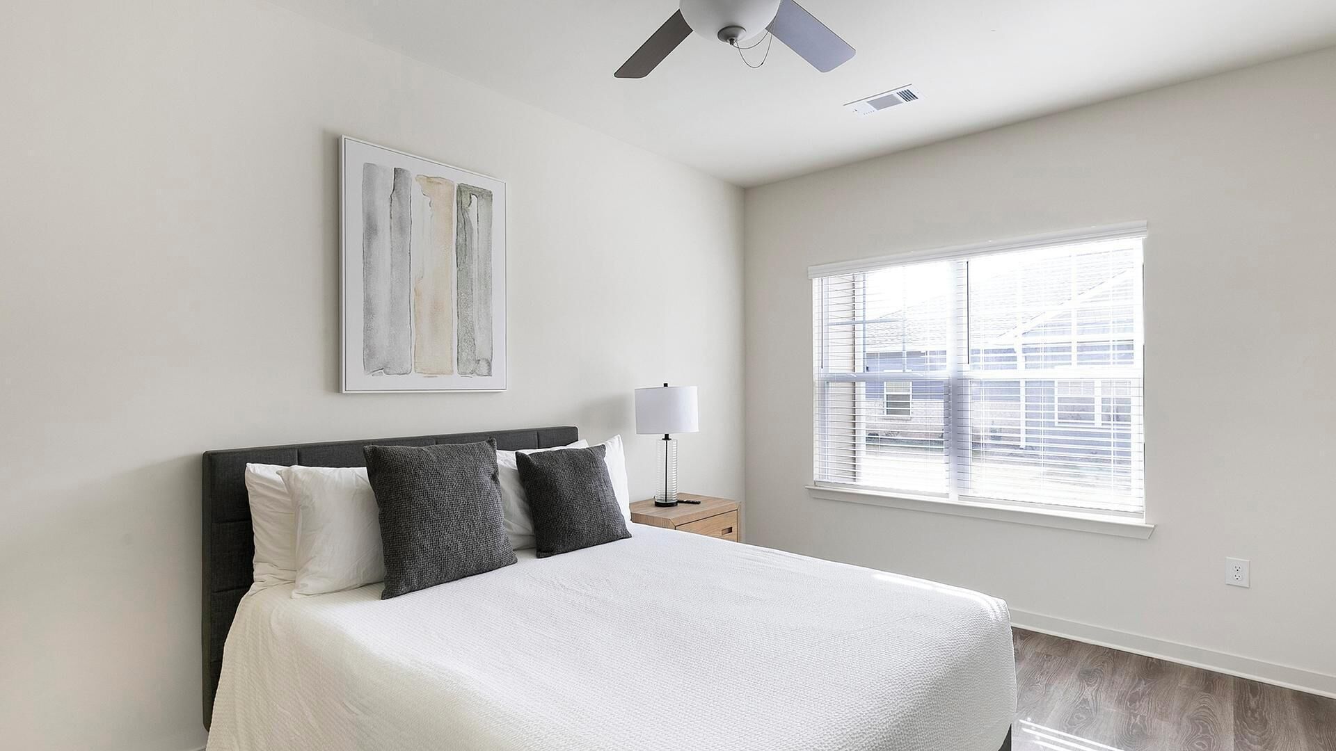 A bedroom with a bed, a nightstand, a lamp, a ceiling fan, and a window with blinds.