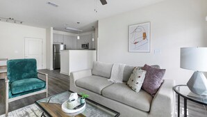 Uma sala de estar moderna com um sofá, uma poltrona verde-água e uma mesa de centro de vidro. Uma cozinha ao fundo, com armários brancos e uma geladeira.