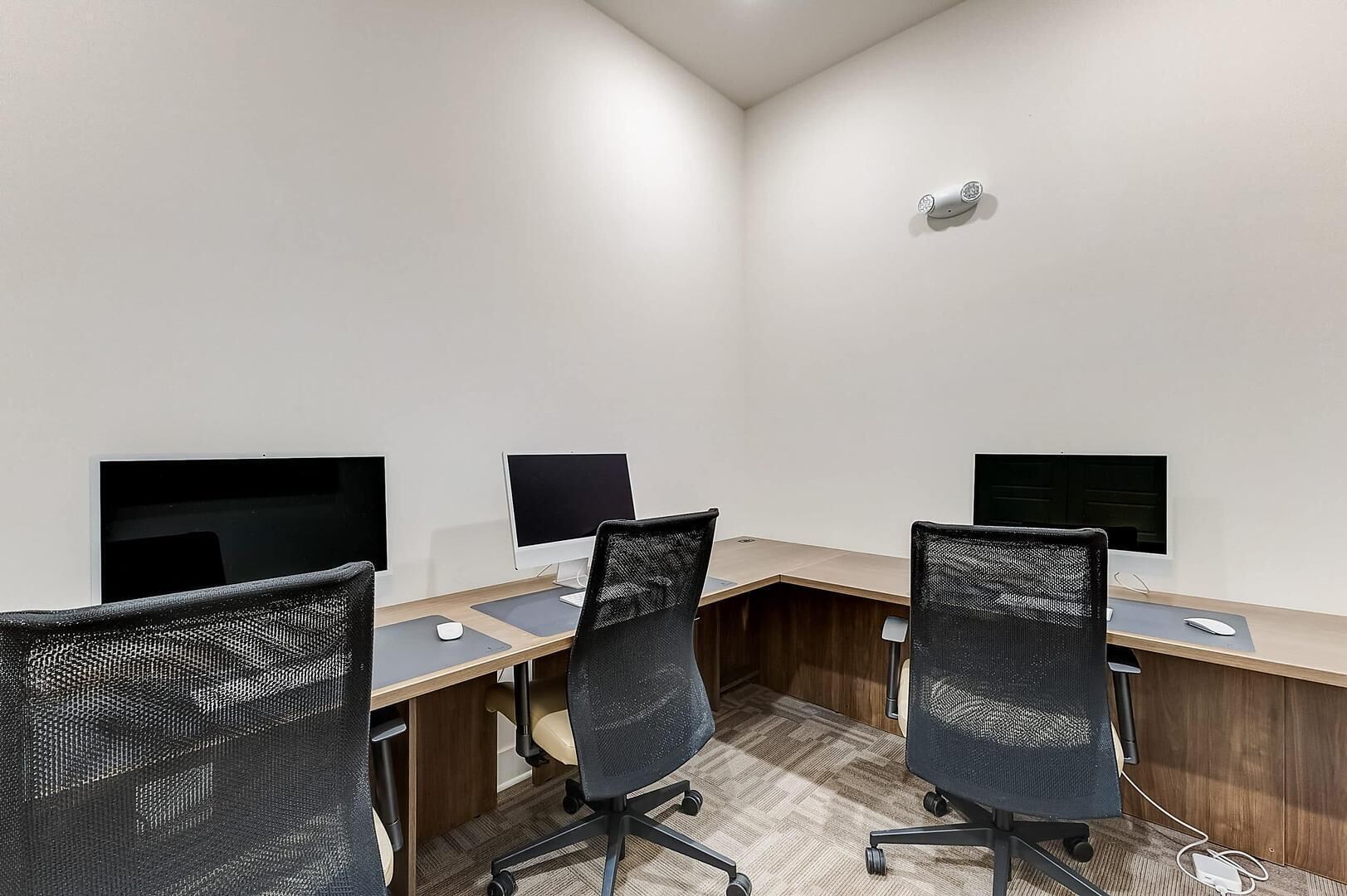 A conference room with three desks, each with a computer monitor, keyboard, and mouse. The room has a white wall, a wooden desk, and black mesh office chairs.