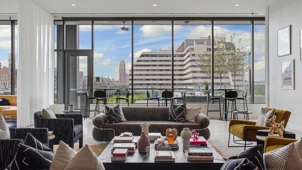 Een moderne woonkamer met een groot raam, een bank, een bijzettafeltje met boeken en uitzicht op een stadsbeeld.
