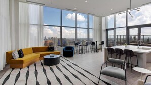 Un salon moderne comprenant un canapé, une table basse et des fauteuils, un coin cuisine avec un bar, et de grandes fenêtres offrant une vue sur la ville.