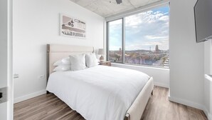 Une chambre avec une grande fenêtre, un lit recouvert de draps blancs, une table de chevet et une vue sur la ville.