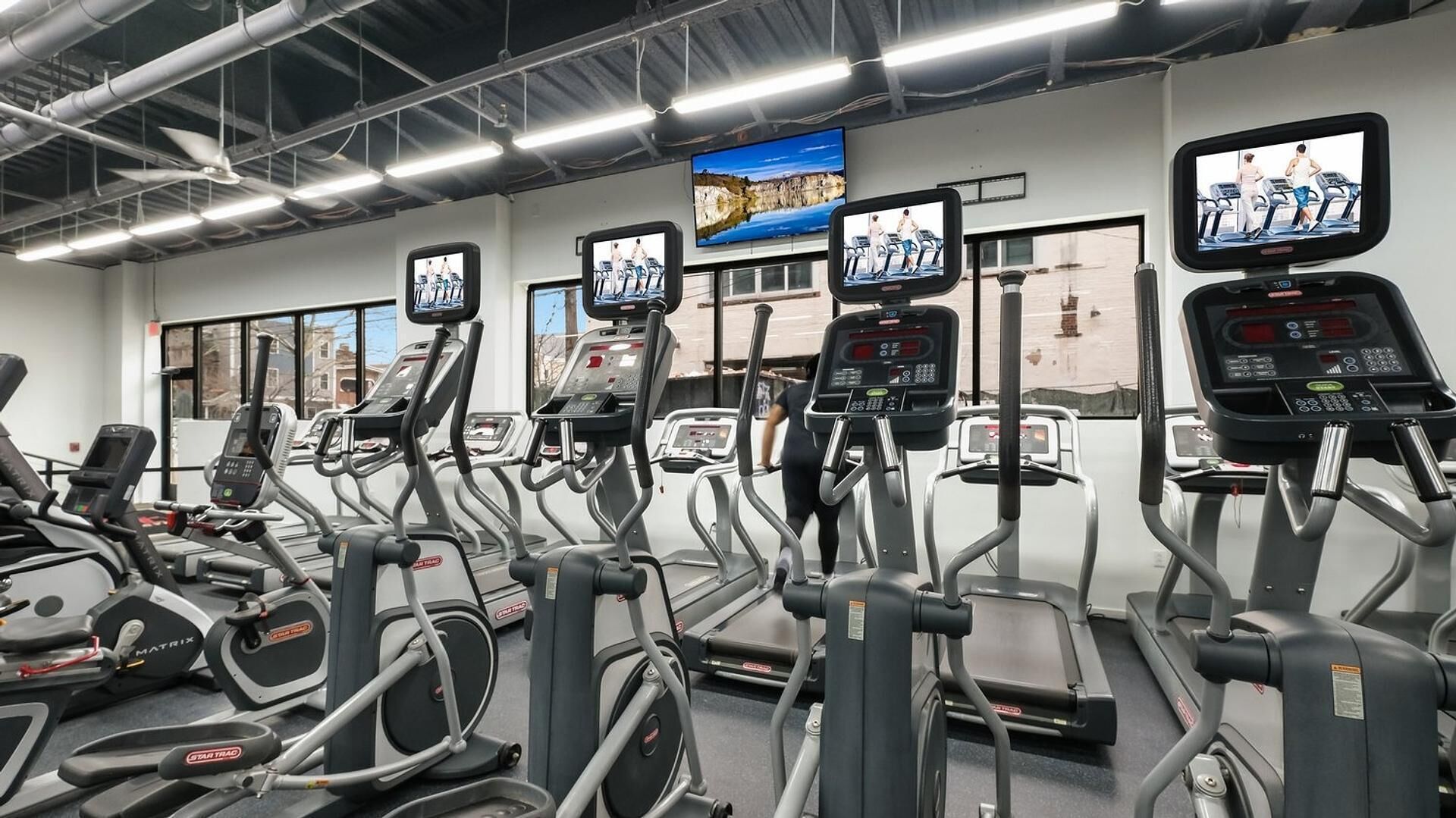 Une salle de sport équipée de plusieurs tapis de course, chacun doté d’un écran numérique et d’un écran affichant des images de personnes en train de courir.