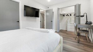 A modern bedroom with a large bed, a flat-screen TV mounted on the wall, a desk with a chair, and a laundry basket. - Landing | Stunning 2BD, Clubhouse, Gym (Philadelphia)