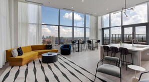 A modern living room with a sofa, coffee table, and chairs, a kitchen area with a bar, and large windows offering a city view.
