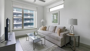 A modern living room with a sofa, coffee table, and lamps. There is a large window with a view of a parking garage. - Landing | Spectacular 2BD, Pool, Gym (Houston)
