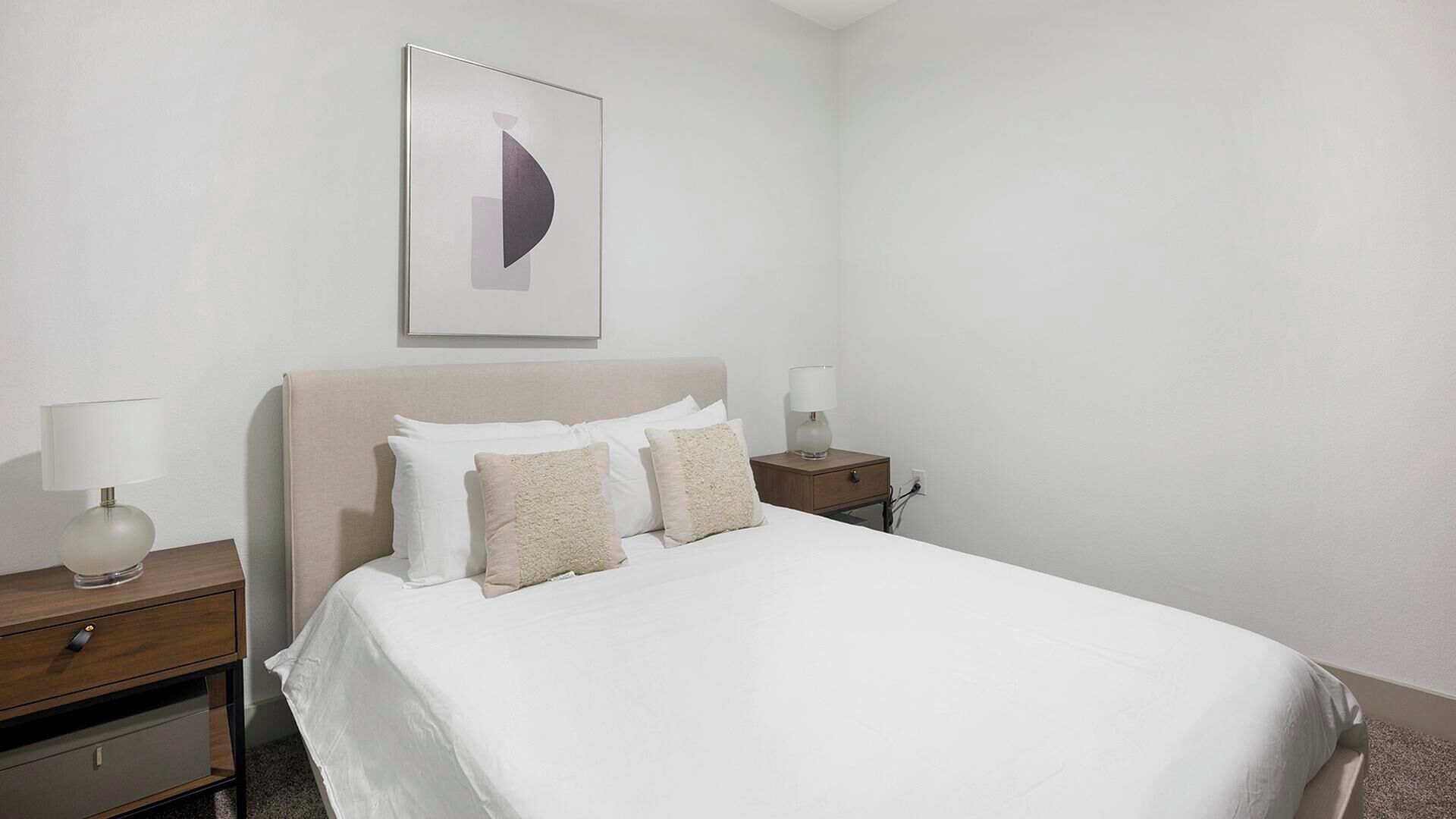 Un lit bien fait, avec du linge de lit blanc et deux oreillers, flanqué de tables de chevet avec des lampes, dans une chambre sobre avec un mur uni et une seule œuvre d’art abstraite au-dessus du lit.
