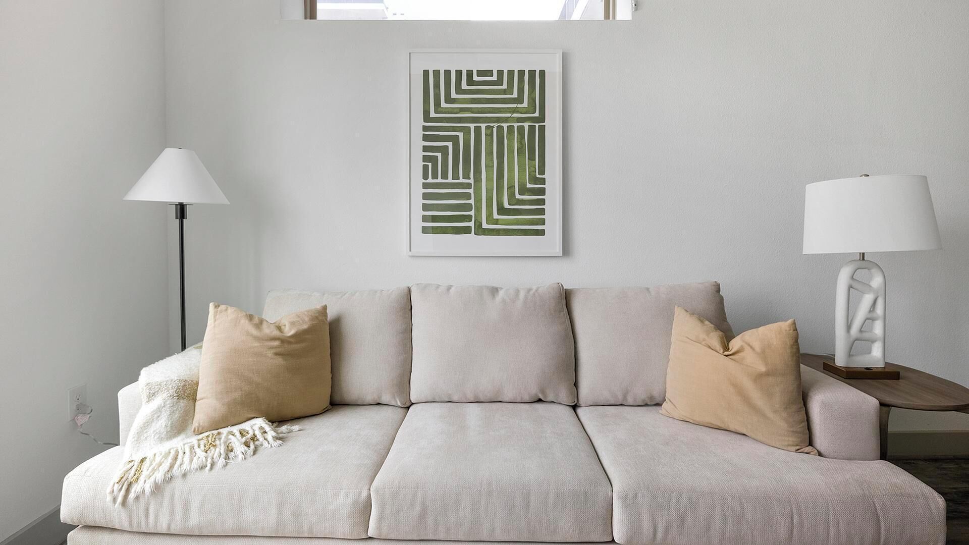 Un sofá de tonos neutros con varios cojines, una lámpara de pie, una lámpara de mesa y una obra de arte abstracta enmarcada en la pared.