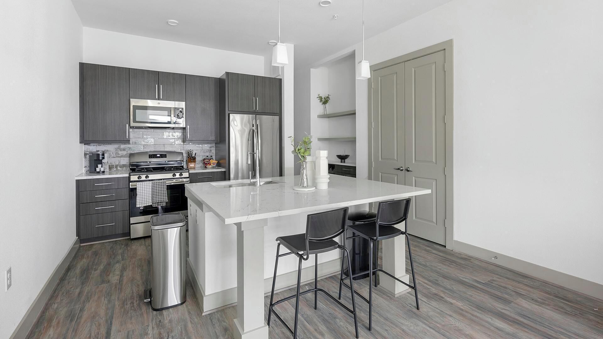 A modern kitchen with grey cabinets, stainless steel appliances, a white island with bar stools, and a grey door.