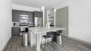 A modern kitchen with grey cabinets, stainless steel appliances, a white island with bar stools, and a grey door. - Landing | Spectacular 2BD, Pool, Gym (Houston)