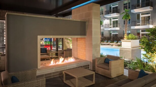 An outdoor lounge area with a fire feature, seating, and a view of a pool and residential buildings. - Landing | Spectacular 2BD, Pool, Gym (Houston)