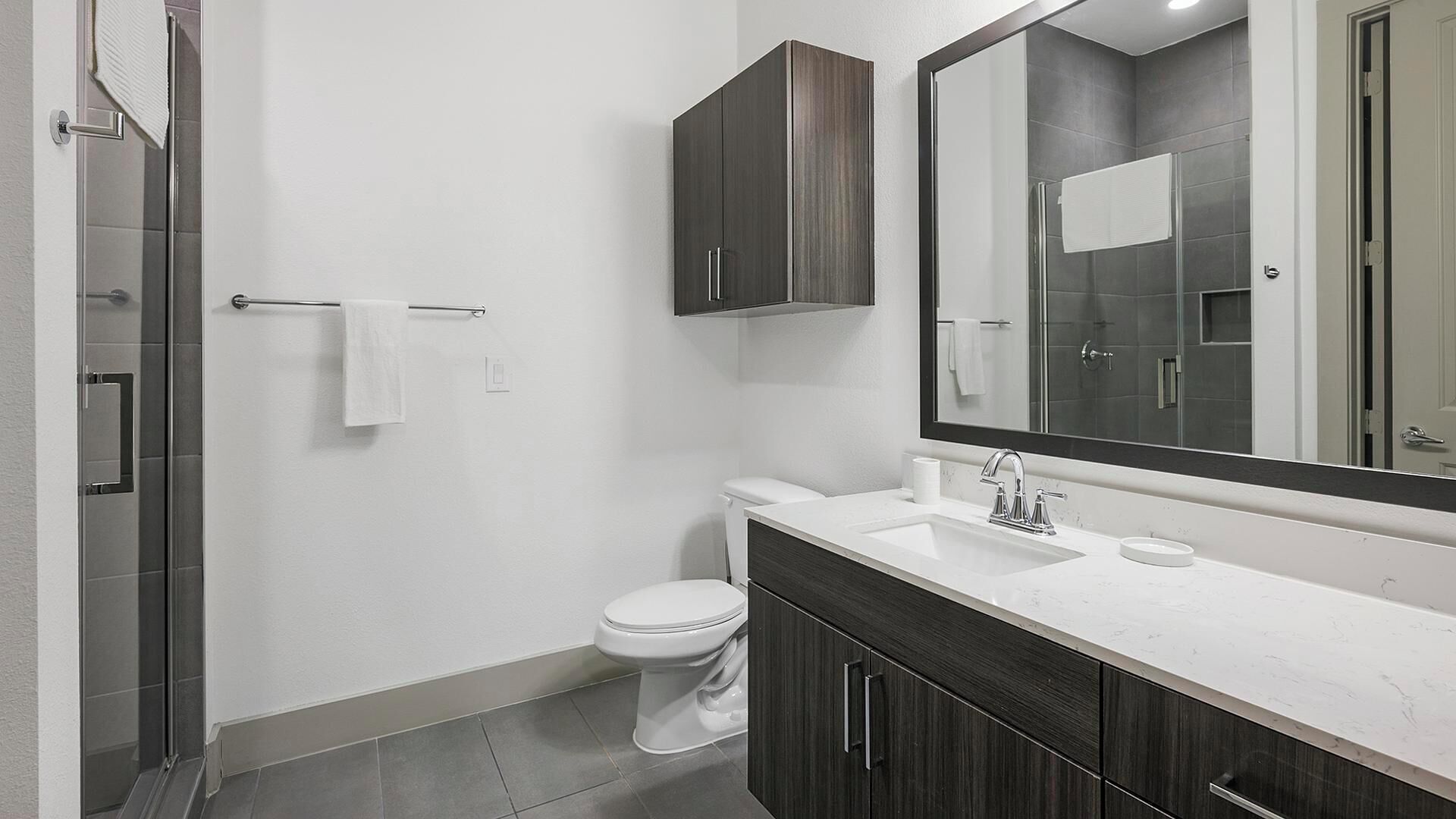 Un baño moderno con inodoro blanco, armarios de madera oscura, un espejo grande y cabina de ducha de vidrio.