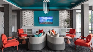 A modern lobby with a teal accent wall, a large flat-screen TV, and a seating area with red and grey chairs and ottomans.