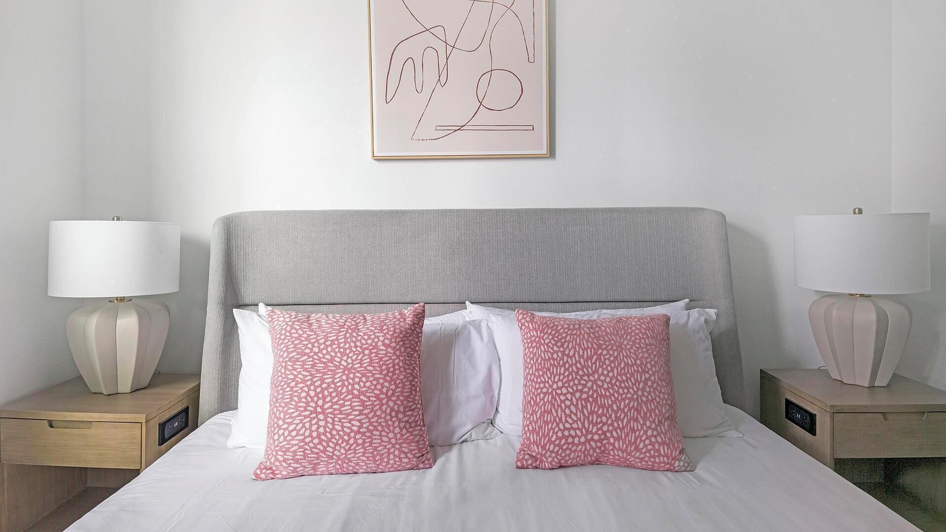 Cama com roupa de cama branca, duas almofadas estampadas em rosa e cabeceira cinza, com duas luminárias de cabeceira em mesas de cabeceira de madeira.