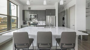 Cozinha moderna com uma ilha grande, armários escuros, eletrodomésticos de aço inoxidável e vista através de amplas janelas.