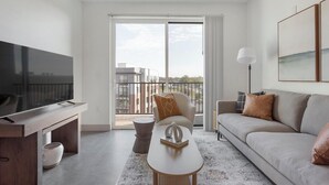 A modern living room with a flat-screen TV, a sofa, a coffee table, and a balcony with a view of buildings. - Landing | Amazing 2BD, Clubhouse, Gym (Richmond)