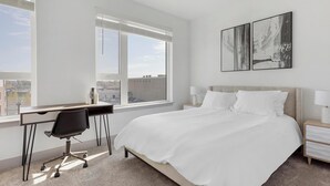 A hotel room with a bed, a desk, and a chair. There are two windows, one with a view of buildings and another with a view of the sky. - Landing | Amazing 2BD, Clubhouse, Gym (Richmond)