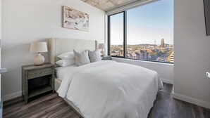 Ein modernes Schlafzimmer mit einem großen Bett, einem Nachttisch, einer Lampe und Blick auf die Stadt durch ein großes Fenster.