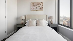 A minimalist bedroom with a large bed, two bedside tables with lamps, a city view through the window, and a painting on the wall.